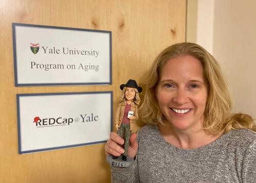 Mary Geda celebrates her award by showing off a bobblehead of her likeness, a gift from the staff at Program on Aging and REDCap@Yale, next to signs that say "Yale University Program on Aging" and "REDCap@Yale".
